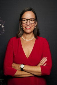 BRAINBOARDS CEO Esther-Mirjam de Boer in einem roten Kleid und Perlenkette mit verschränkten Armen vor dunklem Hintergrund.